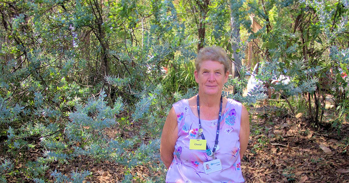 Long-term BlueCare volunteer Cheryl Nott stands amid leafy gardens.