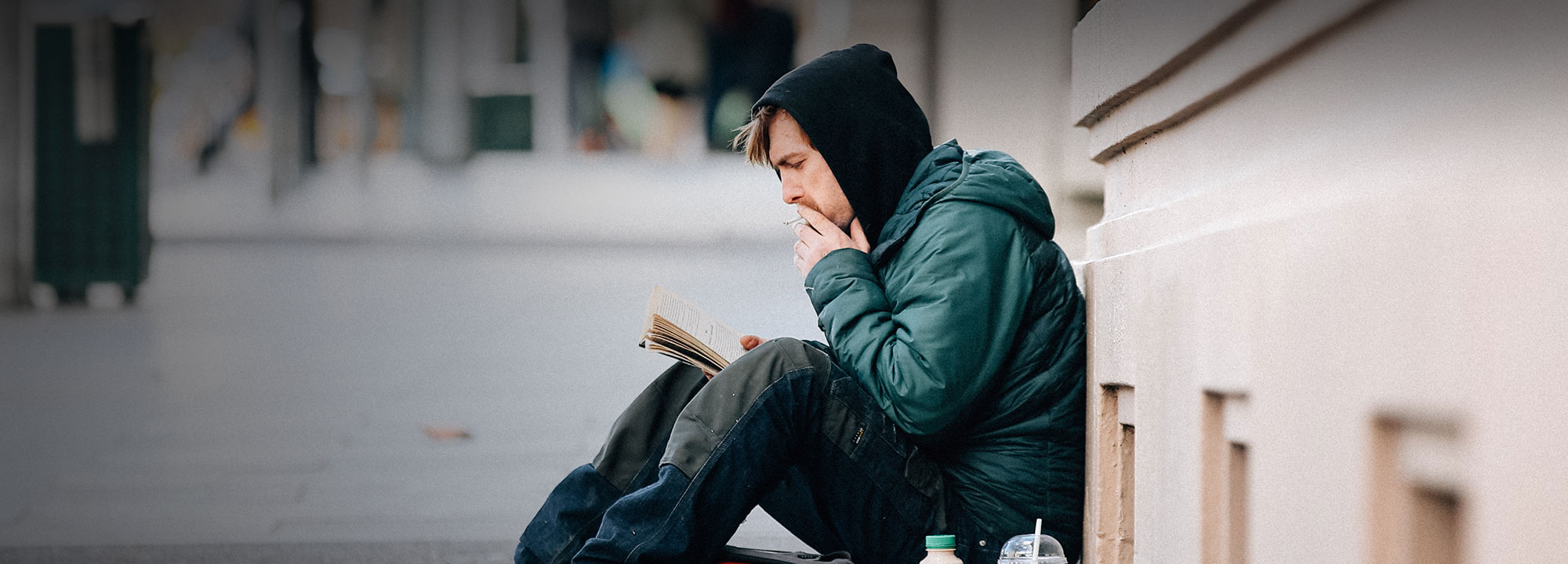 homeless man wearing a hooded jacket reading a book while sitting against a building on a city street
