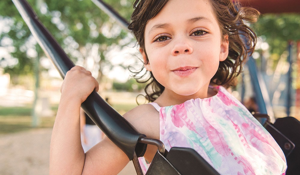 young girl with brown hair smiling at the camera while playing on a swing