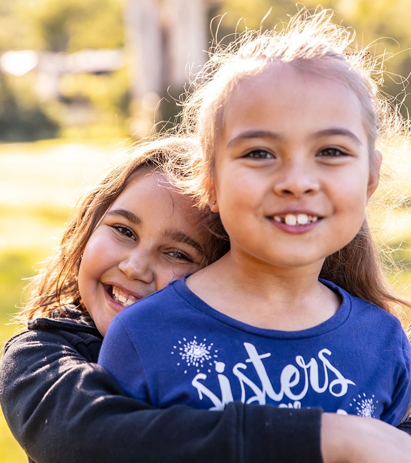 A smiling young girl hugs another smiling young girl in a park.
