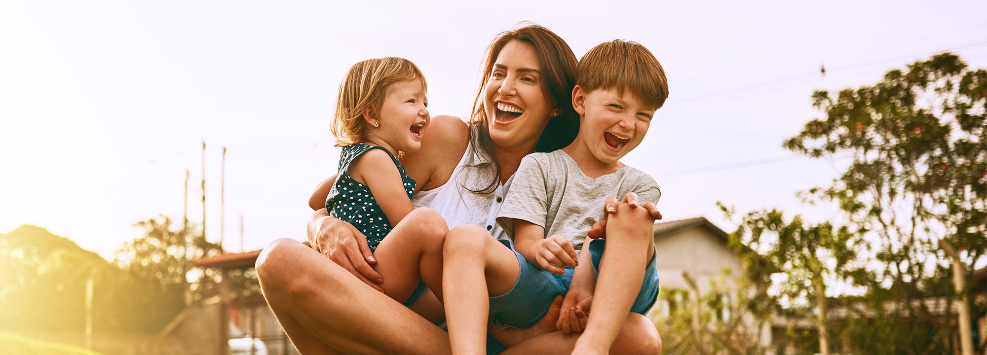 A woman with a young girl and boy in her lap sitting in parklands, laughing and smiling