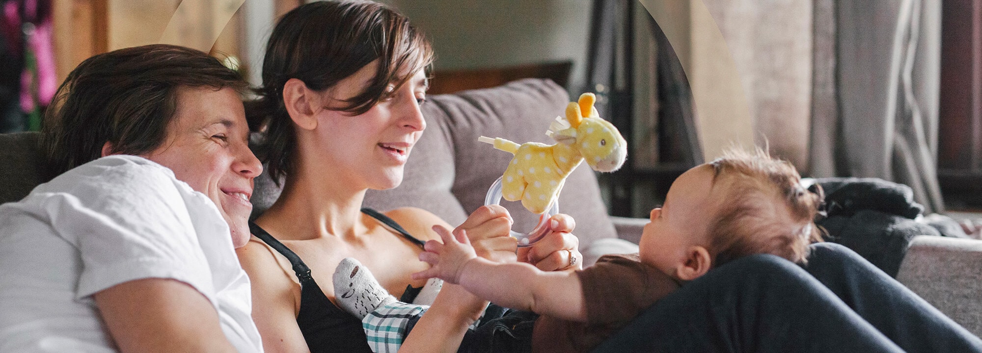 Young mum and dad sit on a couch and play with their baby, who reaches for a yellow toy.